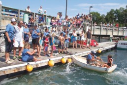 crowd gathered as two people, one blindfolded, row away in a dinghy